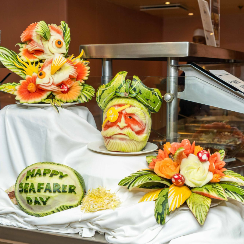 Artistic watermelon carvings of a pirate face, fish, and a 'Happy Seafarer Day' message on a buffet display.