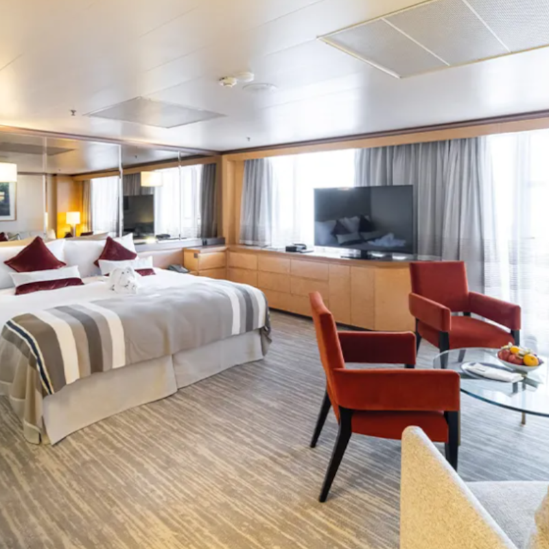 A spacious cruise ship suite featuring a large double bed, a seating area with two red armchairs, a flat-screen TV, and large windows.
