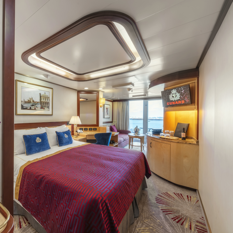 A modern cruise ship suite with a red patterned bed runner, blue accent pillows, and an ocean view through glass doors.