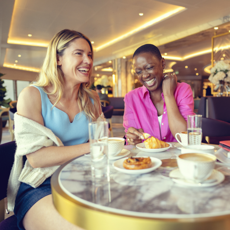 Two women laughing together at a marble table with coffee and pastries inside a cruise ship lounge.