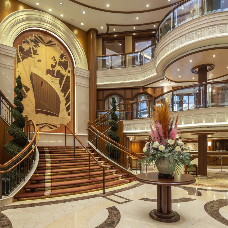 Grand multi-level cruise ship lobby featuring a sweeping grand staircase and a large Art Deco mural of a ship.