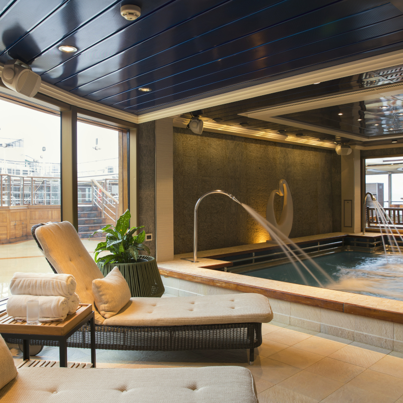 Indoor hydrotherapy pool on a cruise ship with a water fountain feature and cushioned lounge chairs.
