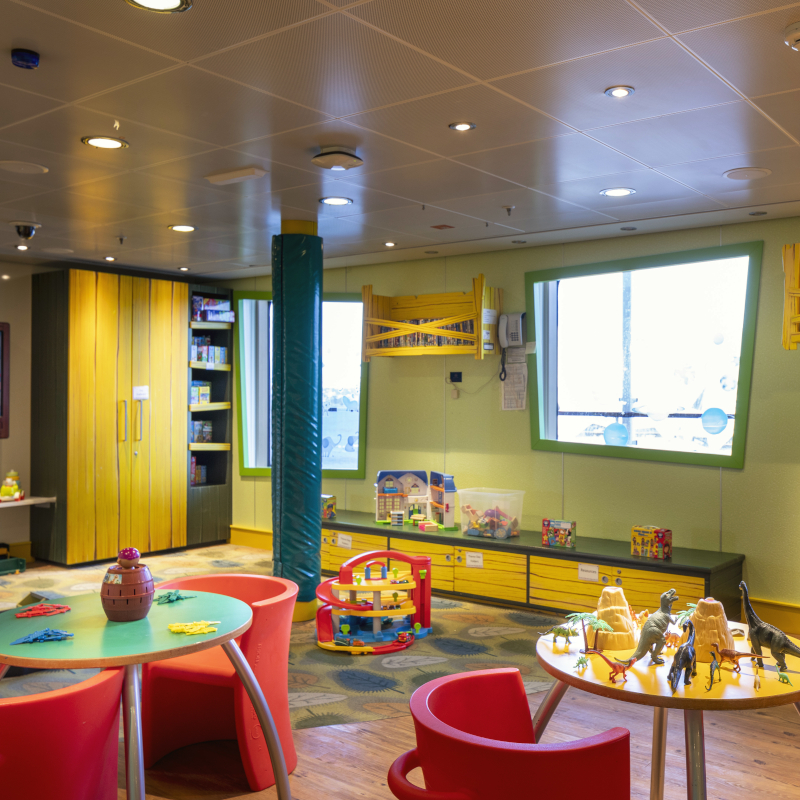 Brightly coloured children's playroom on a ship with toy tables, dinosaur figures, and a yellow storage wall.