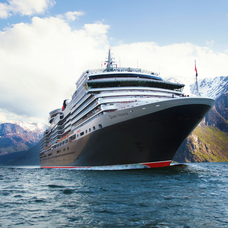 Low-angle view of the Queen Victoria cruise ship sailing through calm water with snow-capped mountains in the background.