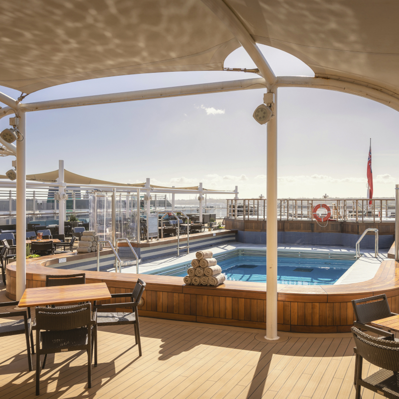 Shaded cruise ship deck pool with wicker tables and chairs.