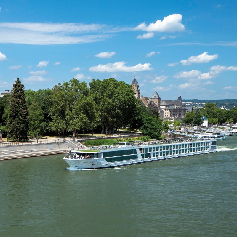 The Emerald Dawn river ship cruising along a wide waterway with lush green trees and historic buildings in the background.