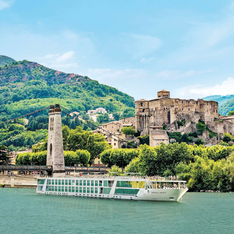 The Emerald Liberté river cruise ship sailing on a river past a historic hilltop castle and a suspension bridge.