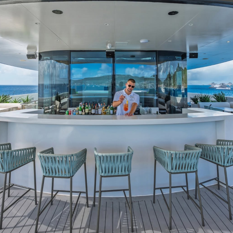 A bartender mixing a cocktail at a sleek, curved white outdoor bar with blue woven bar stools on a cruise ship deck.