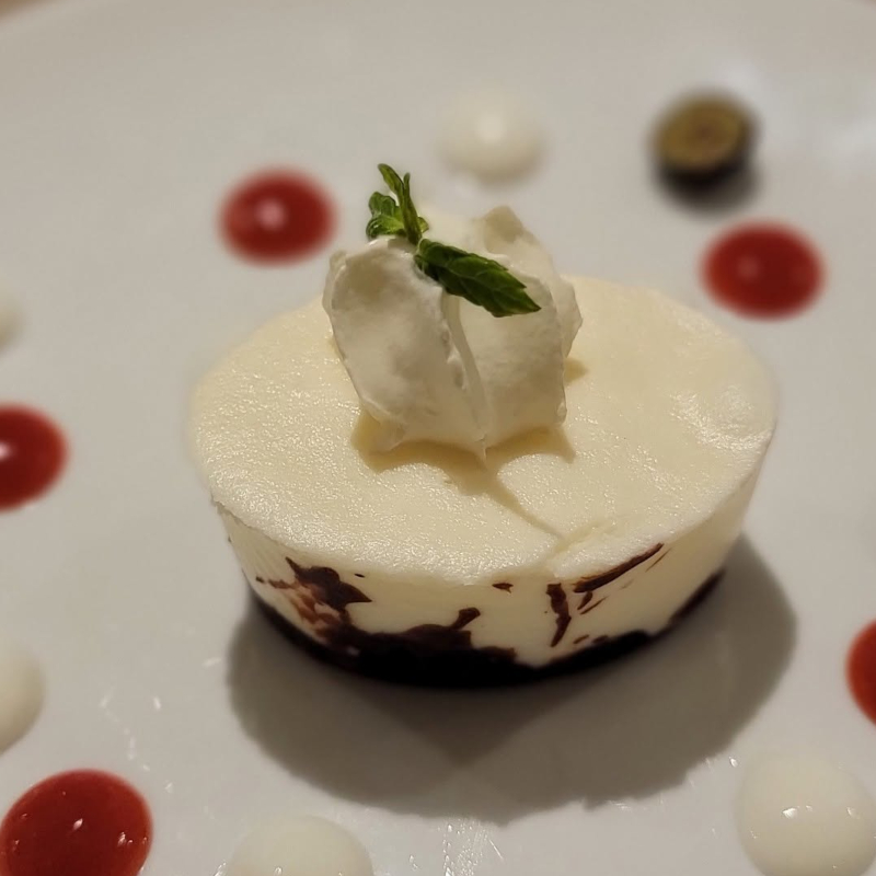 A close-up of a round white chocolate mousse dessert topped with cream and a mint leaf, plated with red and white coulis drops.