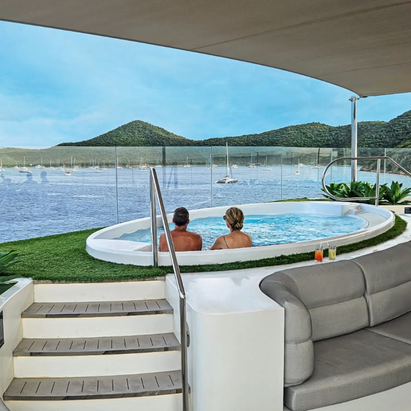 A couple relaxing in an outdoor hot tub on a cruise ship deck, looking out at a bay filled with yachts and green hills.