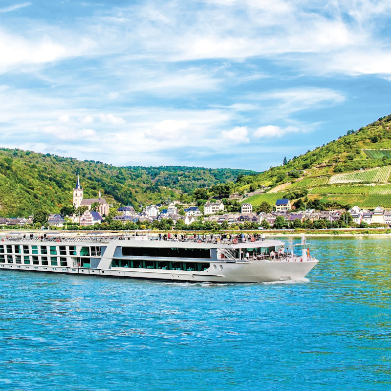An Emerald river cruise ship sailing along a blue river past a hillside town with terraced vineyards.