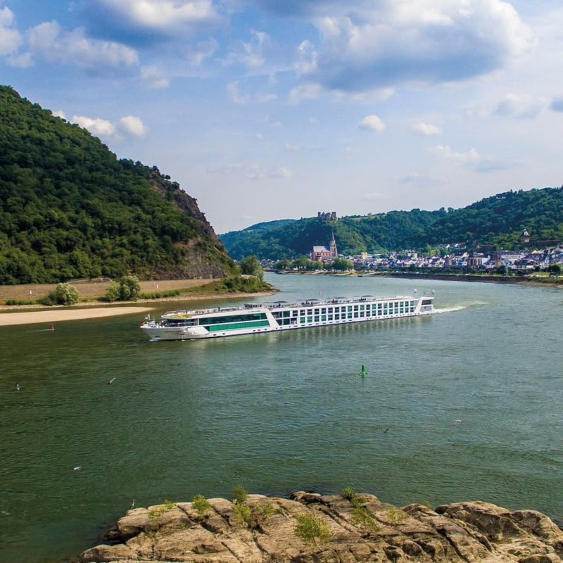 An elevated view of a long white river cruise ship navigating a wide bend in a river flanked by lush green hills.