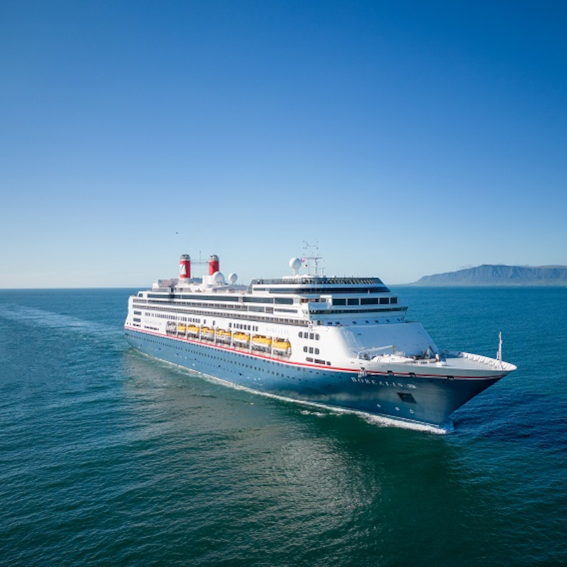 The Borealis cruise ship sailing through deep blue open waters under a clear sky, with mountains visible on the distant horizon.