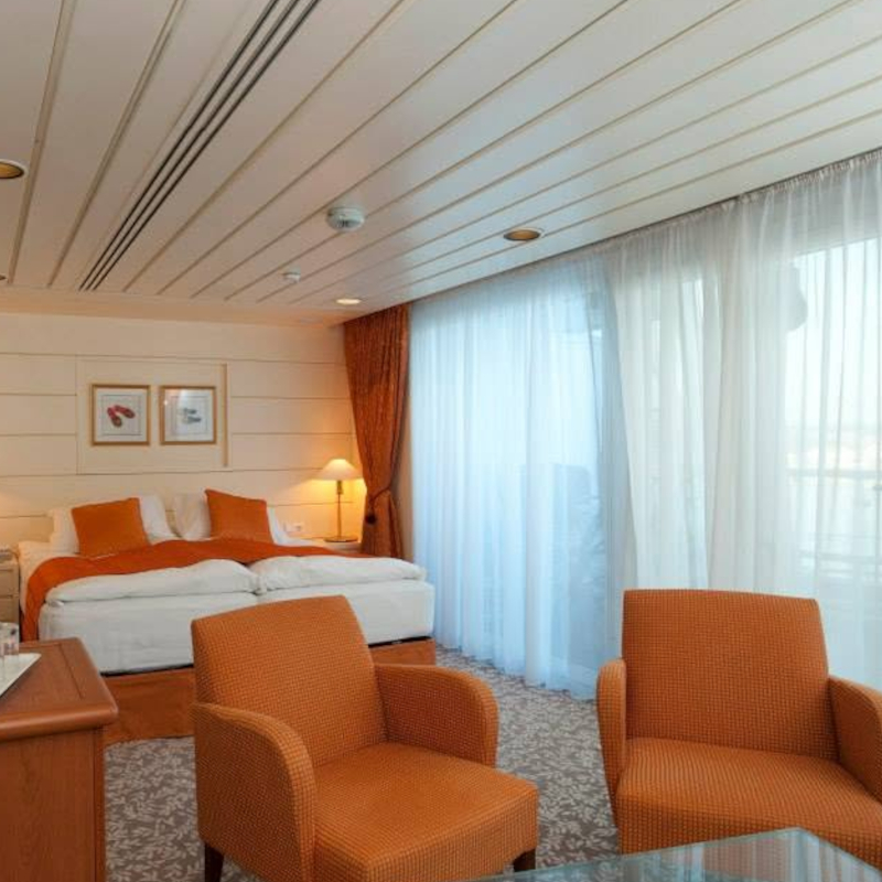 A spacious cruise ship cabin with a bed, two orange armchairs, a wall-mounted TV, and sheer curtains leading to a balcony.