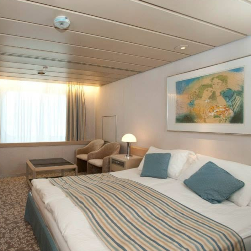 A spacious superior outside cabin with a large double bed, blue striped linens, a navy blue sofa, and a wide window letting in natural light.
