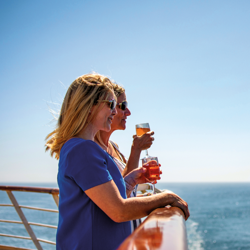Two women standing at the wooden railing of a cruise ship, holding drinks and looking out at a bright blue sea.
