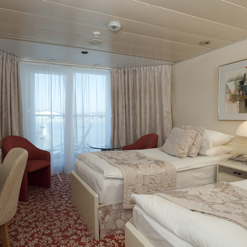A cruise ship twin balcony cabin featuring two single beds with beige floral runners, red accent armchairs, and floor-to-ceiling glass doors leading to a balcony.