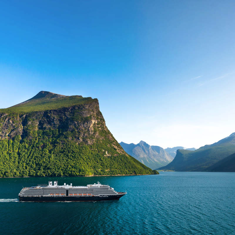 Cruise ship sailing along blue waters past lush green mountains in a Norwegian fjord under a clear blue sky.