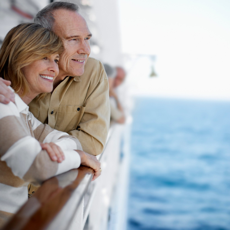 Smiling senior couple leaning against a cruise ship railing while looking out at the open ocean.