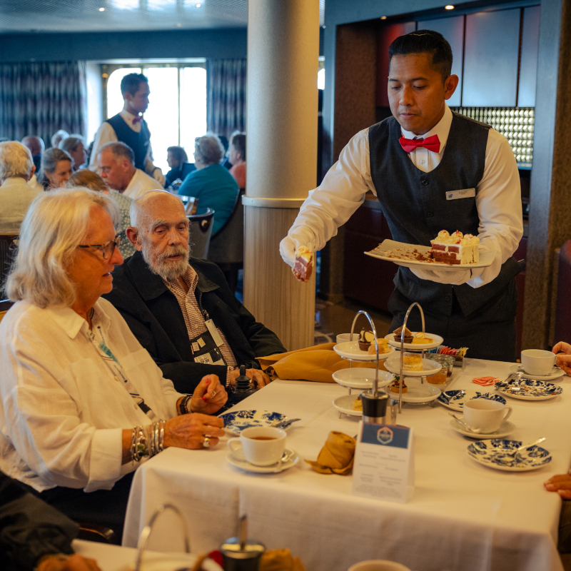 Waiter in a vest and red bow tie serving a slice of cake to guests during afternoon tea on a cruise ship.