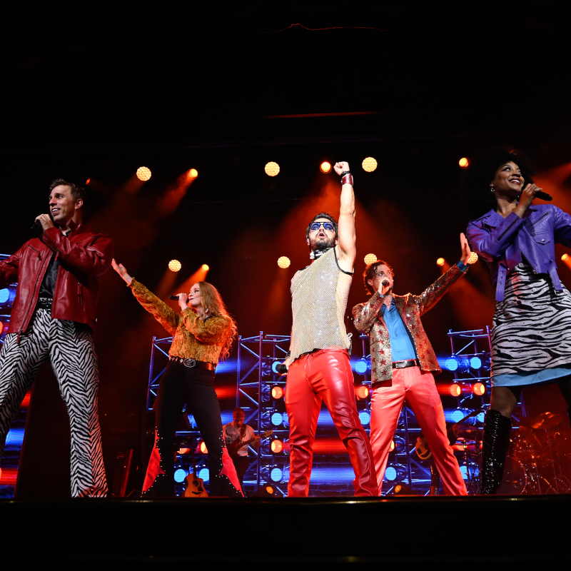 Five performers in colourful, retro 1970s outfits singing and dancing on a brightly lit stage.