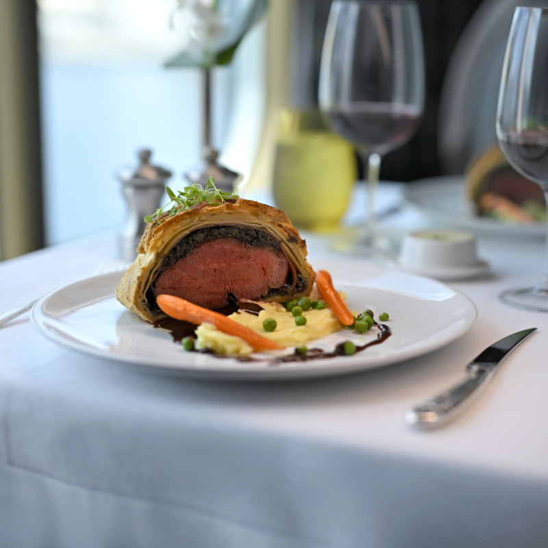 Gourmet Beef Wellington served with mashed potatoes, baby carrots, and peas on a white plate on white tablecloth.
