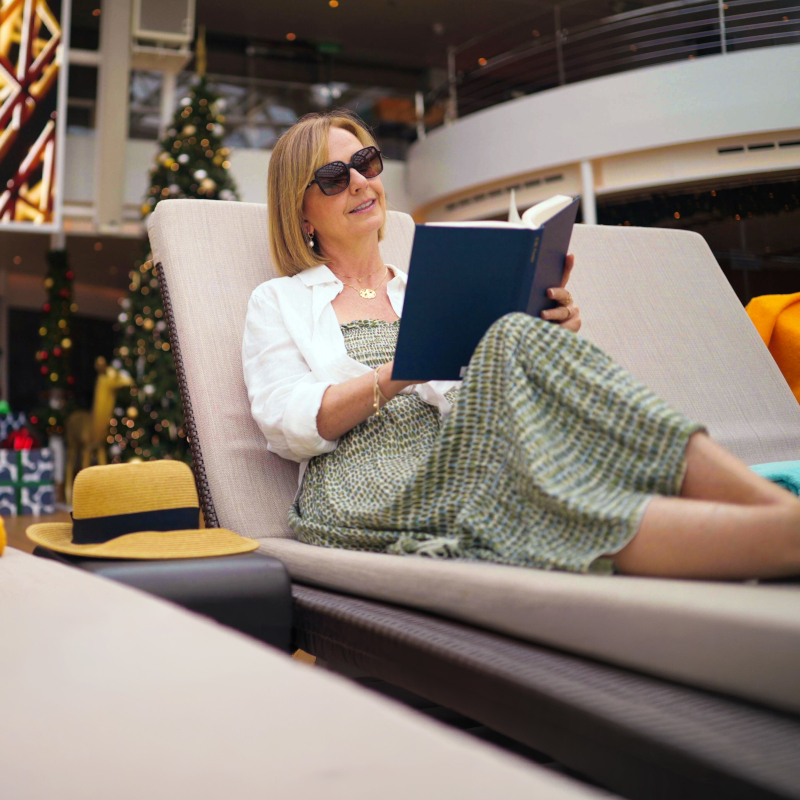 Woman wearing sunglasses relaxing on a lounge chair on a cruise deck while reading a book.