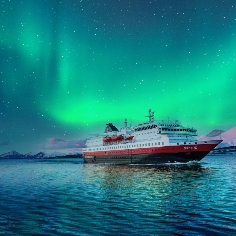 Hurtigruten ship Nordlys sailing at night beneath brilliant green Northern Lights and a starry sky.
