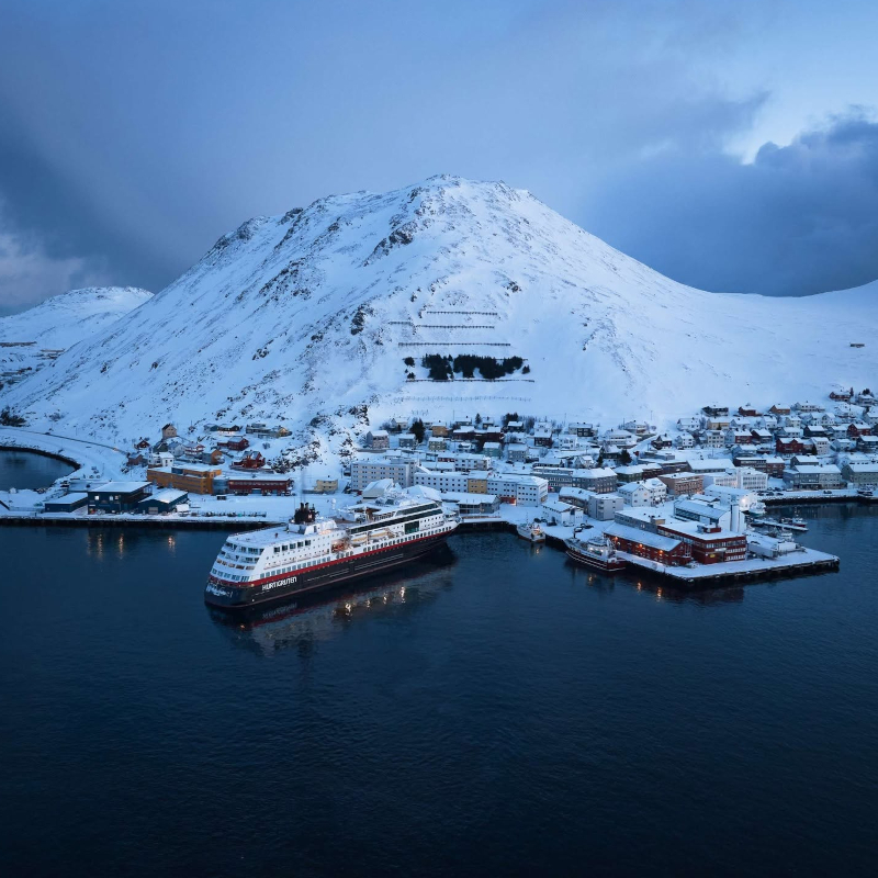Aerial view of a Hurtigruten ship docked at a snow-covered coastal town with a large mountain behind it.