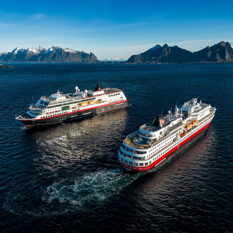 Aerial view of two Hurtigruten ships cruising side-by-side on deep blue water near snow-capped mountains.