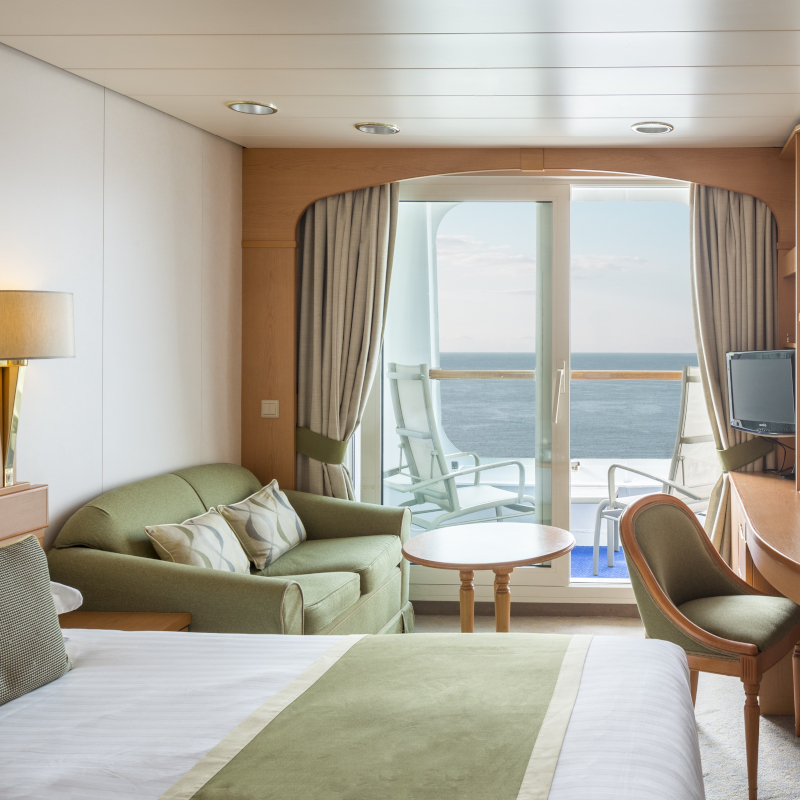 A modern cruise ship balcony cabin featuring a bed, sofa, and glass sliding door opening to an ocean view.