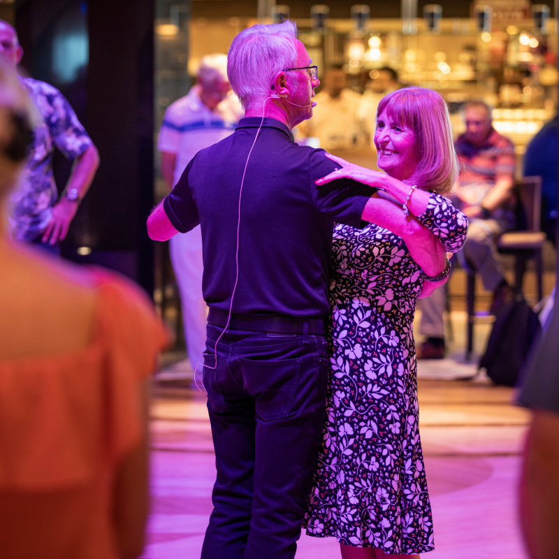 A smiling couple enjoying a ballroom dance class under vibrant purple lighting on board.