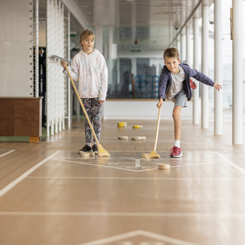 Two children playing shuffleboard together on the outdoor wooden deck of a cruise ship.