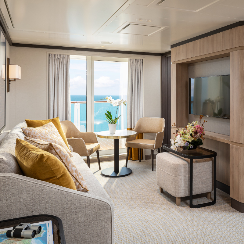 A modern cruise ship suite living room with a sofa, chairs, TV, and balcony ocean view.