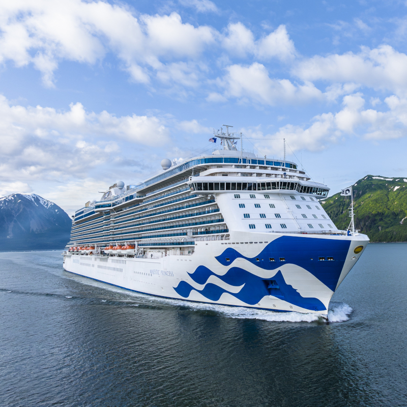 Front-three-quarter view of the Majestic Princess cruise ship navigating water with mountains in the background.