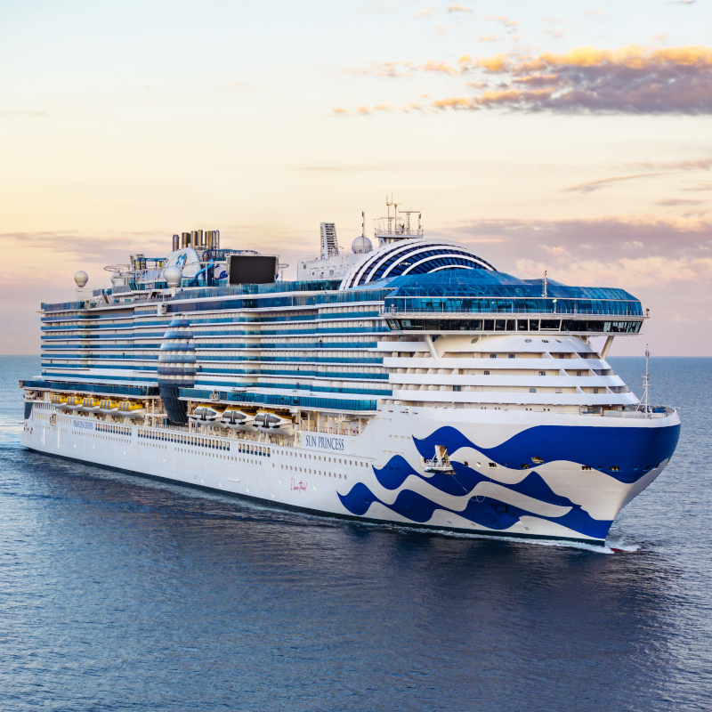 Bow side view of the Sun Princess cruise ship sailing on blue ocean waters under a warm twilight sky.