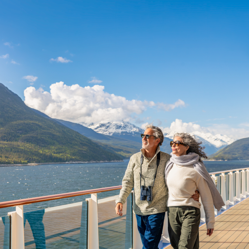A couple walking along an open promenade deck with snow-capped mountains in the background.